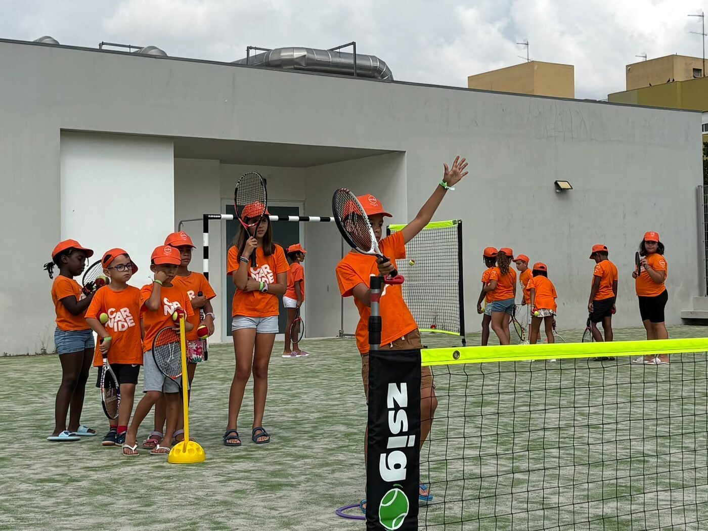 Rapariga a acenar num campo de ténis ao ar livre, com o grupo de participantes de laranja