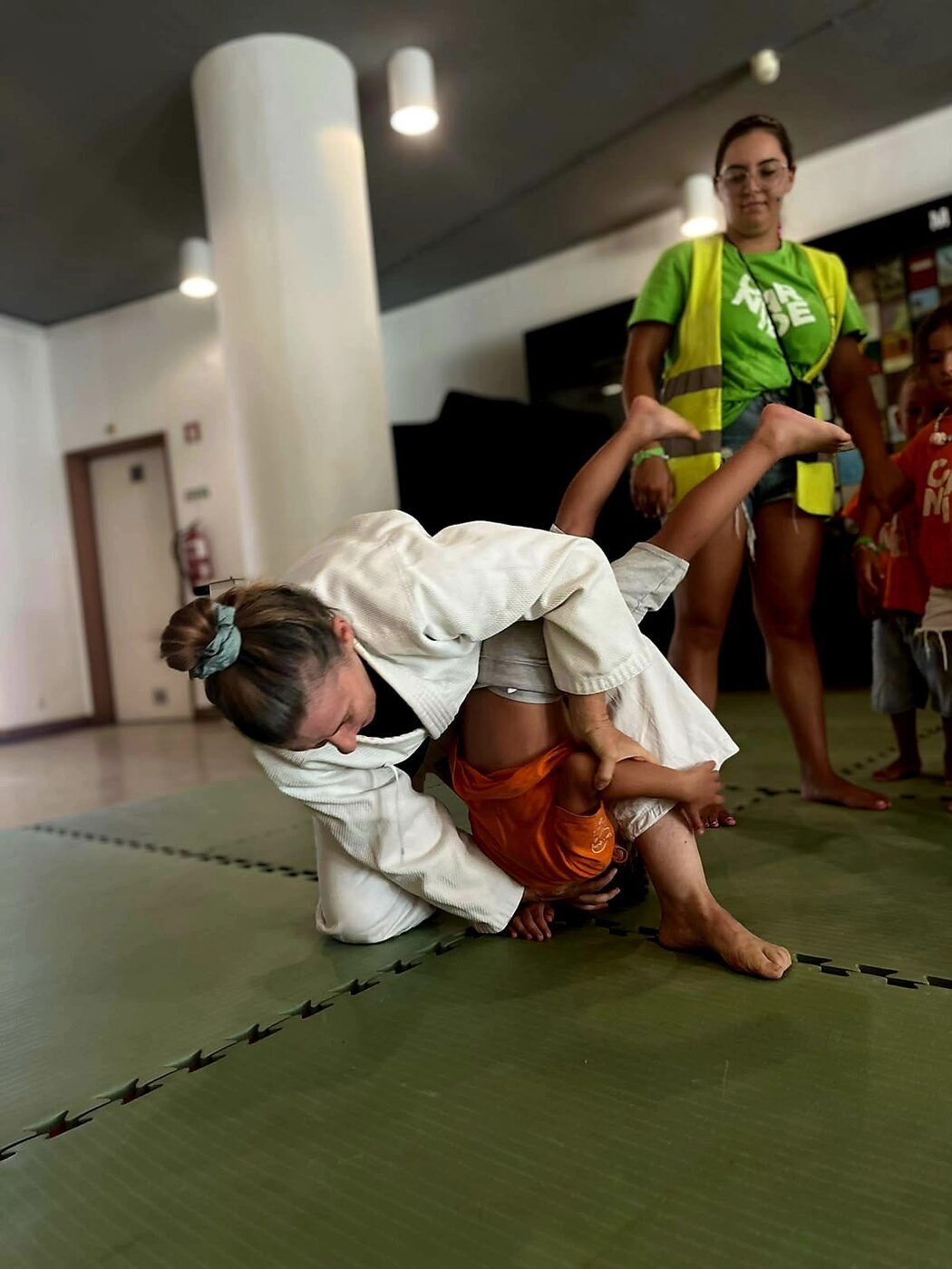 Criança a fazer uma cambalhota num tapete de judo, com o monitor