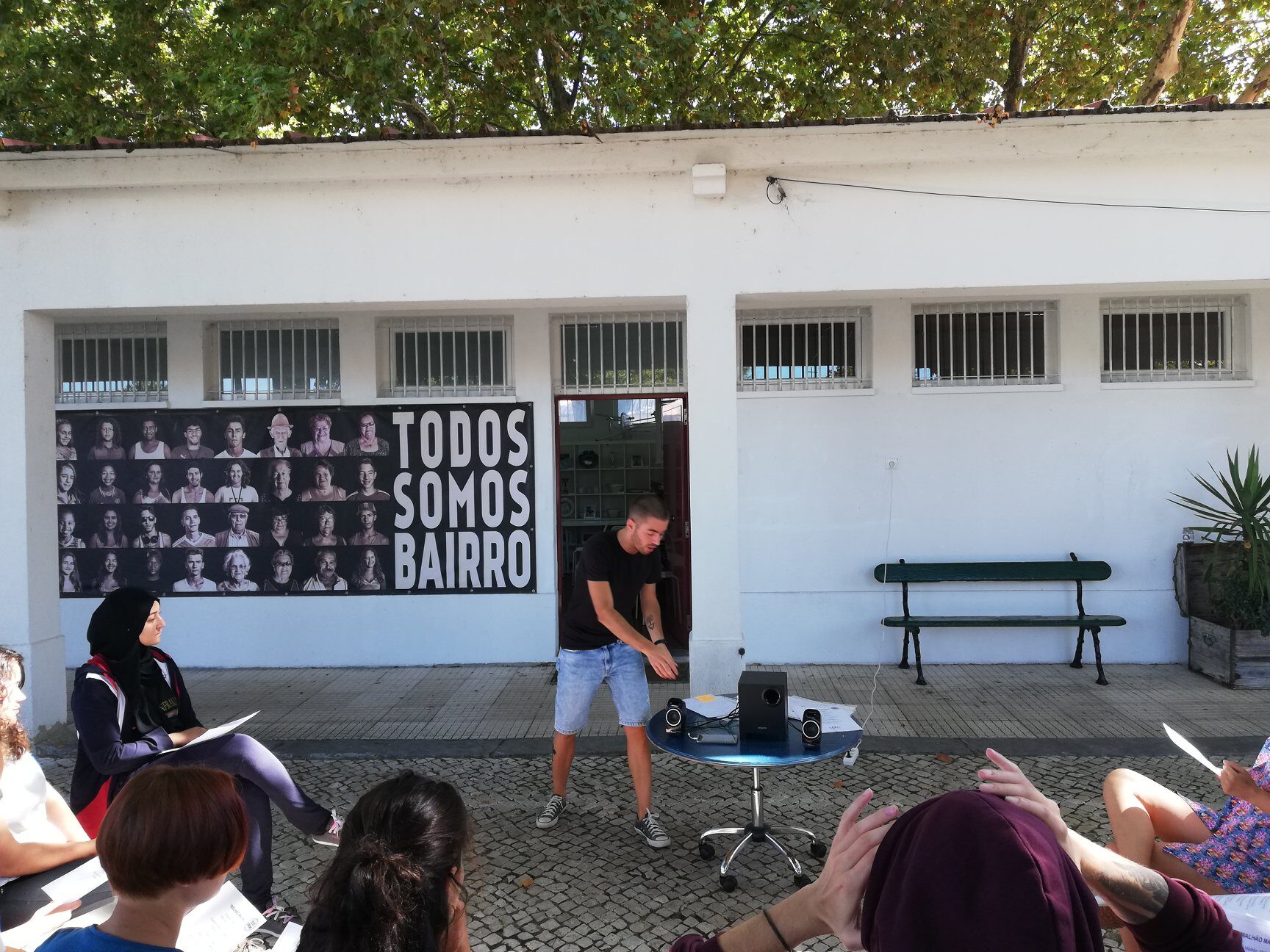 Exterior do Espaço Comunitário do Bairro Padre Cruz com o mural 'Todos Somos Bairro' e comunidade reunida